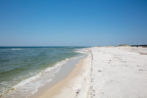 A serene beach scene with white sand, gentle waves, and a clear blue sky.