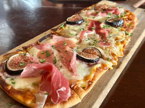 A flatbread pizza topped with cheese, figs, prosciutto, and herbs, served on a wooden board.