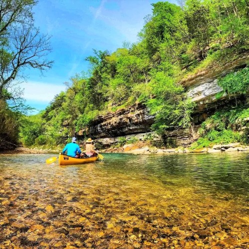 Two people in a yellow canoe paddle along a clear river surrounded by lush green trees and rocky cliffs.