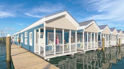 Colorful waterfront houses on stilts with porches, connected by a wooden walkway, under a bright blue sky.