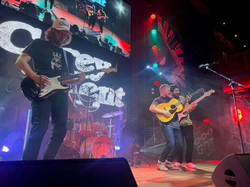 A band performs on stage with guitars, colorful lights, and a large screen in the background.