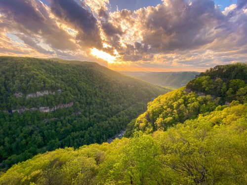 A scenic view of lush green hills and valleys under a dramatic sky with clouds and sunlight breaking through.