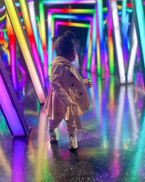 A child in a light pink coat stands in a colorful, illuminated tunnel with vibrant neon lights.