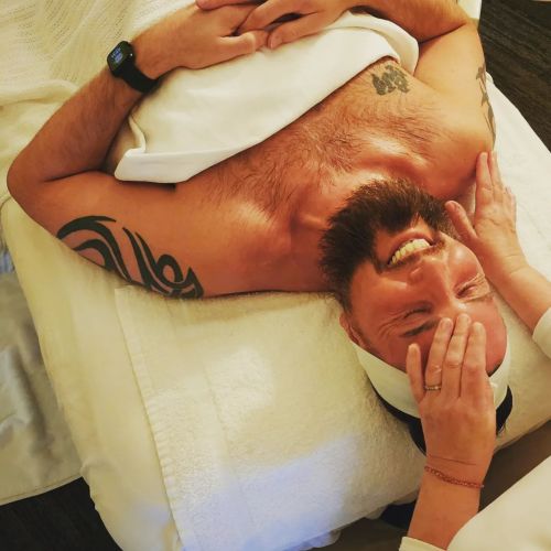 A man with tattoos lies on a massage table, smiling as a therapist gently massages his forehead.