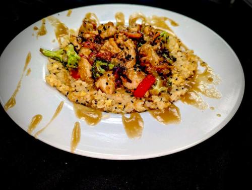 A plate of stir-fried chicken with vegetables served over rice, drizzled with sauce and garnished with sesame seeds.