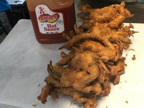 A stack of fried frog legs next to a bottle of Texas Pete hot sauce on a white paper surface.