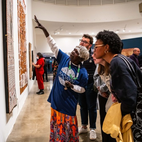A group of people engages with an art exhibit, as a guide points out details on the wall.