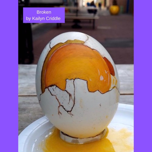 A large, cracked egg sculpture with a vibrant orange yolk, titled "Broken" by Kailyn Criddle, displayed outdoors.