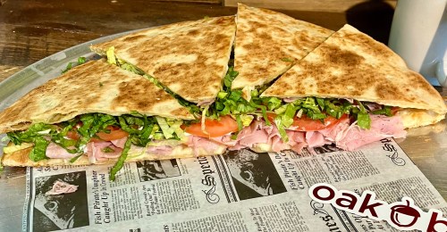 A large, sliced quesadilla filled with ham, lettuce, and tomatoes, served on a metal tray with newspaper lining.