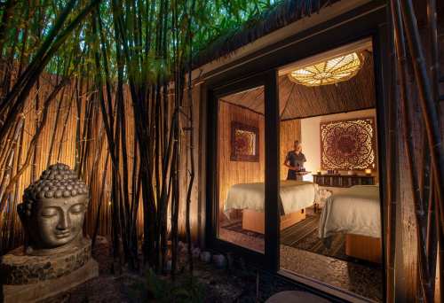 A serene spa room with bamboo surroundings, featuring a Buddha statue and a person preparing for a massage.