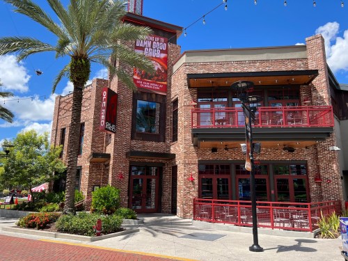A brick building with red accents, featuring outdoor seating and palm trees, under a blue sky with scattered clouds.