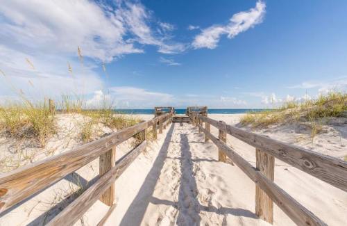 A wooden walkway leads through sandy dunes to a serene beach under a blue sky with scattered clouds.