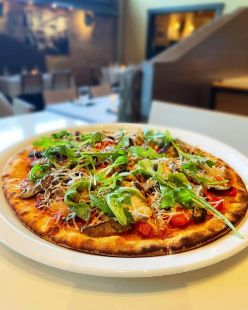 A freshly baked pizza topped with greens, tomatoes, and cheese, served on a white plate in a restaurant setting.