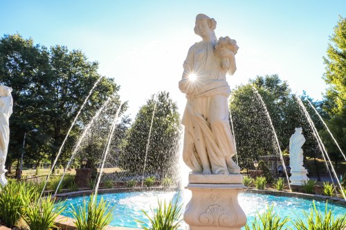 A white statue holding flowers stands in a fountain, with sunlight shining through the water jets and greenery surrounding it.