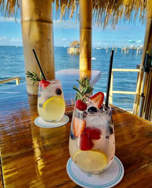 Two refreshing cocktails with fruit garnishes sit on a wooden table by the ocean, with a straw and a scenic view in the background.