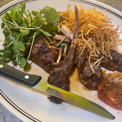 A plate of grilled lamb chops topped with crispy onions, served with greens and a slice of baked tomato.