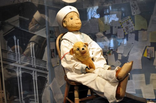 A vintage doll in a sailor outfit sits on a chair, holding a small stuffed animal, surrounded by notes and a reflective background.