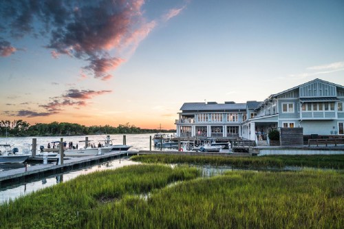 A serene waterfront scene at sunset, featuring a restaurant by the water and boats docked near lush green marshland.
