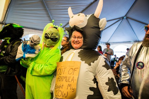 A group of people in costumes, including an alien and a cow, with a sign reading "OUT OF FORMULA - Bought My COW."