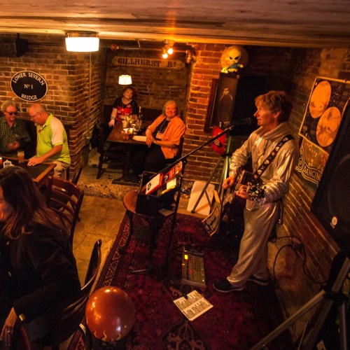 A musician in a silver outfit performs with a guitar in a cozy bar, while patrons enjoy the atmosphere.
