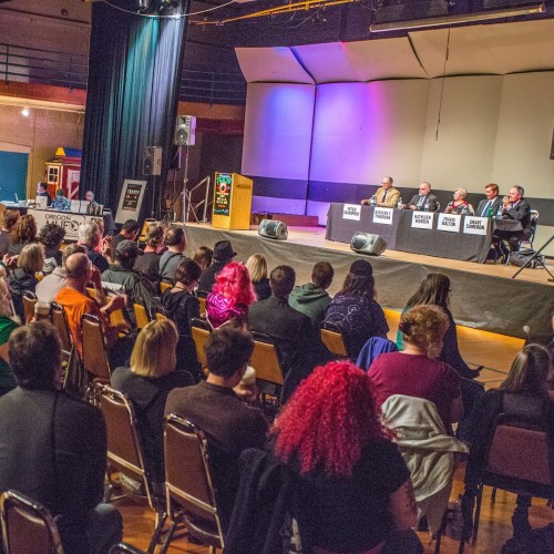 A crowd listens to a panel of five speakers on stage at an event in a large auditorium.