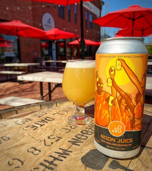 A can of Noon Juice beer next to a glass of the same beer, set on a wooden table with red umbrellas in the background.
