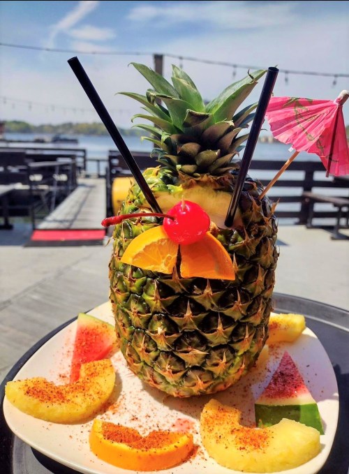 A pineapple cocktail garnished with fruit, straws, and a small umbrella, served on a plate with watermelon slices.
