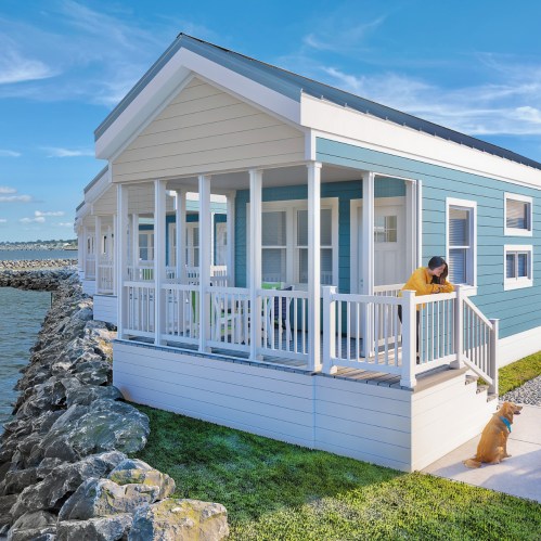 A modern blue beach house with a porch, overlooking water, featuring a person leaning on the railing and a dog sitting nearby.