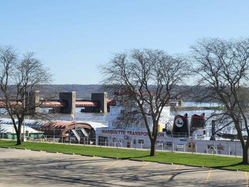 A riverboat docked near a shoreline, with trees and a clear blue sky in the background.