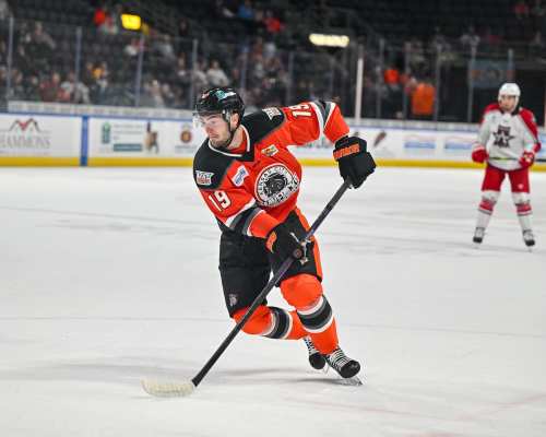 A hockey player in an orange jersey skates with a puck during a game, with another player in the background.