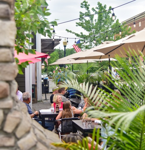 Outdoor dining scene with people at tables under umbrellas, surrounded by greenery and string lights.