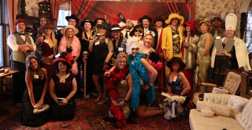 A large group of people in costumes and hats posing together in a decorated room with a vintage vibe.