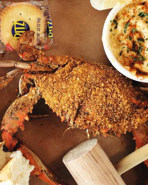 A seasoned crab on a plate with a side of grits, a cracker, and a wooden mallet for cracking the shell.