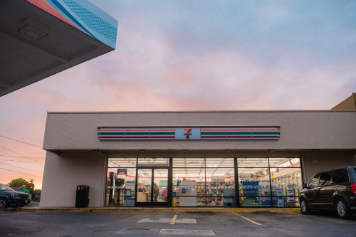 A 7-Eleven store at dusk, featuring large windows and a colorful sign, with a parking lot in front.