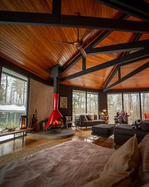 Cozy living room with wooden ceiling, modern furniture, and a red fireplace, surrounded by large windows and nature views.