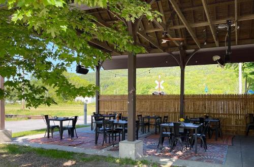 Outdoor dining area with black tables and chairs, surrounded by greenery and a wooden fence, under a shaded roof.