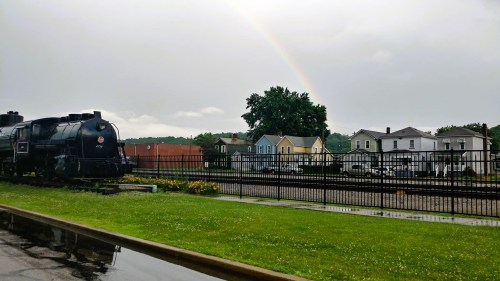 A vintage steam train beside a grassy area, with colorful houses and a rainbow in the background under a cloudy sky.