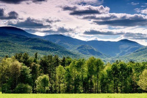 Lush green trees in the foreground with rolling mountains and a cloudy sky in the background.