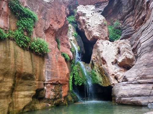 A serene waterfall cascades over rocks into a tranquil pool, surrounded by lush greenery and rugged cliffs.