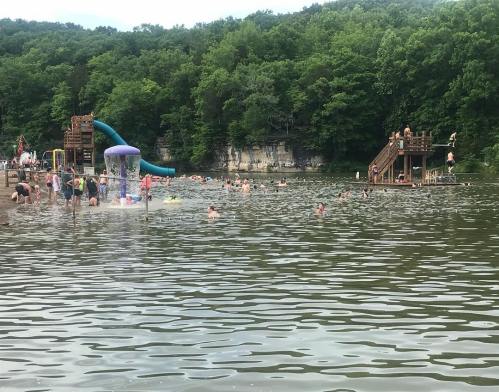 A busy lake scene with people swimming, a water slide, and a wooden platform surrounded by trees.