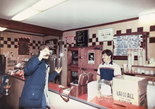 A man gestures excitedly at a counter while a smiling employee stands behind it in a vintage snack shop.