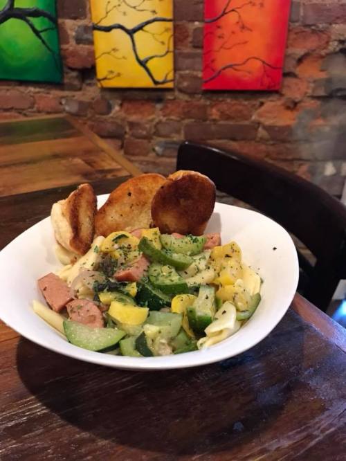 A bowl of pasta with vegetables and ham, served with two fried dough pieces, against a colorful brick wall backdrop.