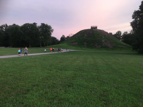 People walking on a path near a grassy area and a large mound under a colorful sky at dusk.