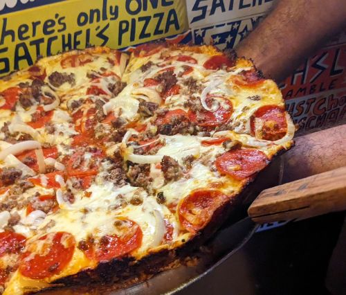 A person holds a large pizza topped with pepperoni, sausage, onions, and cheese, against a colorful background.