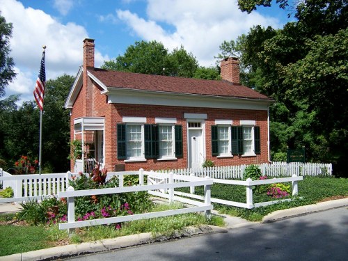 A charming brick house with white trim, surrounded by a white picket fence and colorful flowers, under a blue sky.
