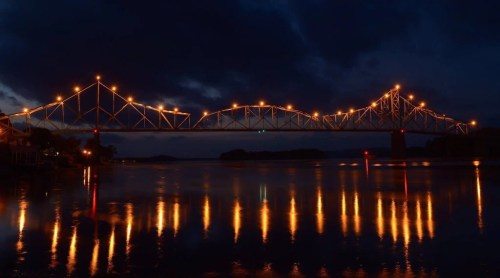 A illuminated bridge spans a calm river at night, reflecting warm lights on the water's surface under a dark sky.