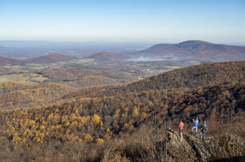 Two hikers stand on a rocky outcrop, overlooking a colorful autumn landscape of rolling hills and valleys.