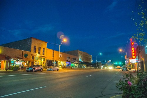 A quiet street scene at dusk, featuring shops and cars under a blue sky with streetlights illuminating the road.