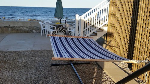 A striped hammock is set up by the seaside, with a patio and stairs in the background.
