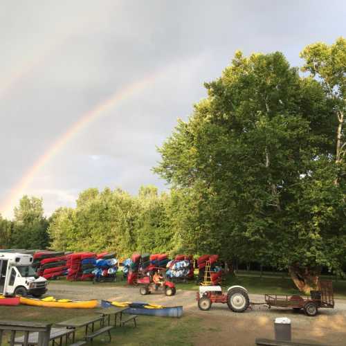 A vibrant rainbow arcs over a campsite with kayaks, a large tree, and parked vehicles in a lush green setting.